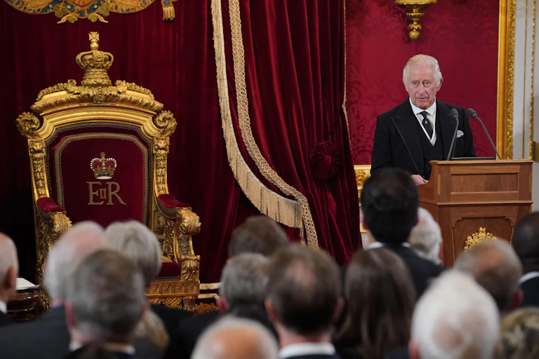 The king stands at a lecturn besides a throne.