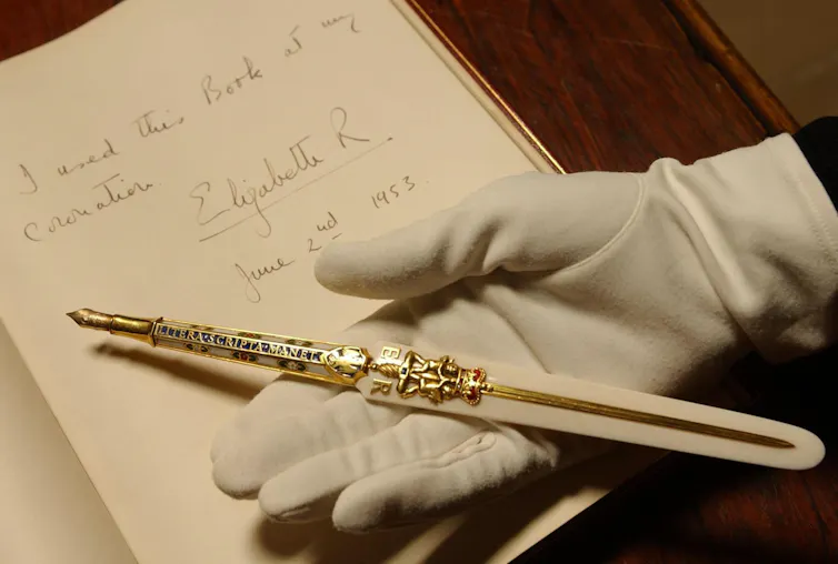 A gloved hand holds a gold pen atop an ecru page of handwriting.