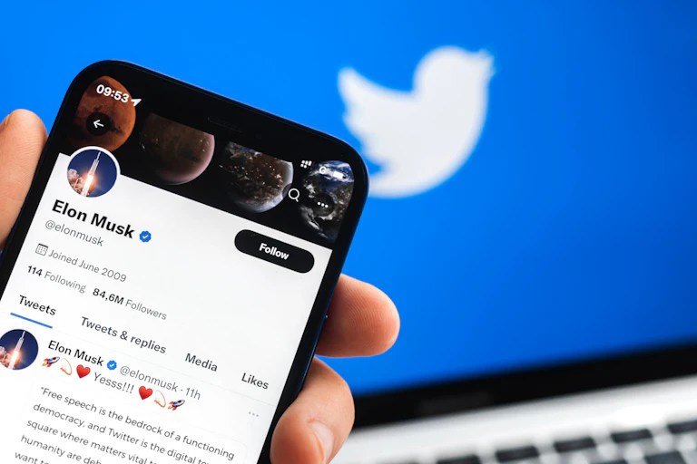 A hand holds an iPhone showing Elon Musk's twitter profile on the screen.