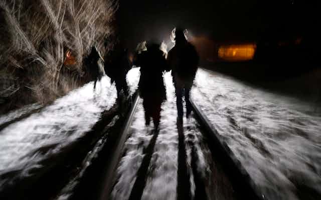 A blurred photo shows the shadows of migrants as they walk along a railroad track in the darkness.