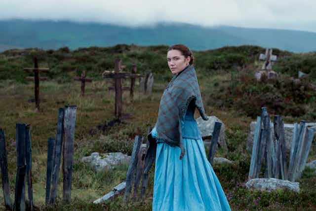 Florence Pugh looks to camera against the backdrop of an Irish graveyard, full of wooden crosses. She wears a long blue dress and a blue plaid shawl round her shoulders. 