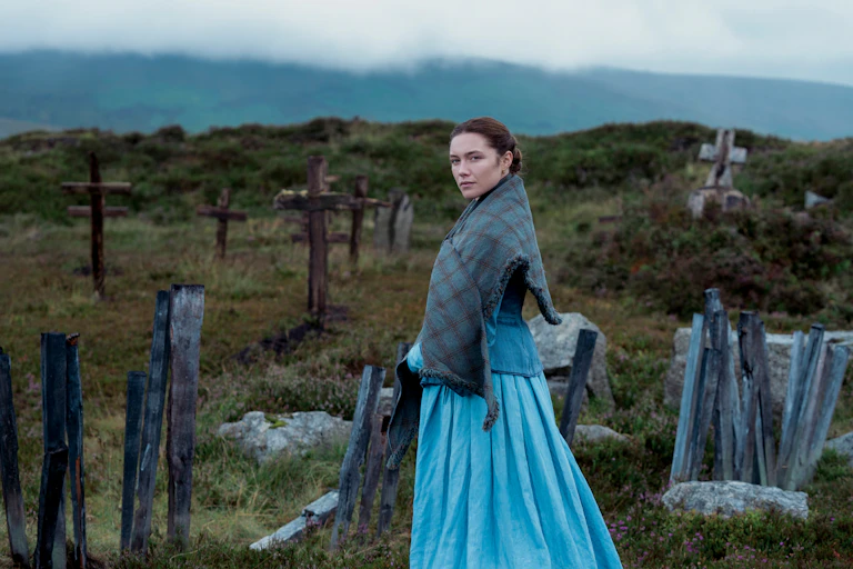 Florence Pugh looks to camera against the backdrop of an Irish graveyard, full of wooden crosses. She wears a long blue dress and a blue plaid shawl round her shoulders.