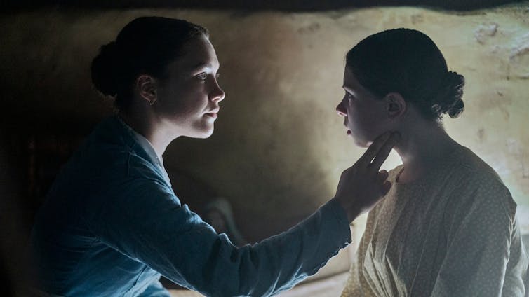 Both actresses are pictured in profile, in low candle light. Florence Pugh reaches out to take Lila Lord Cassidy's pulse.