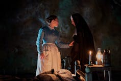 In a dark candlelit room, Florence Pugh (in a blue dress and white apron) puts her hand on a nun's arm.