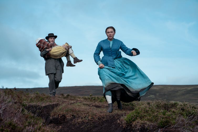 Tom Burke carries Kíla Lord Cassidy in his arms across a rugged landscape, while Florence Pugh in blue dress runs off ahead.