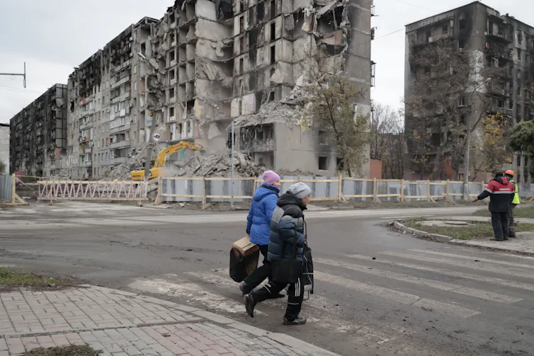 Two women in heavy coats walk past several destroyed and burnt-out apartment buildings.