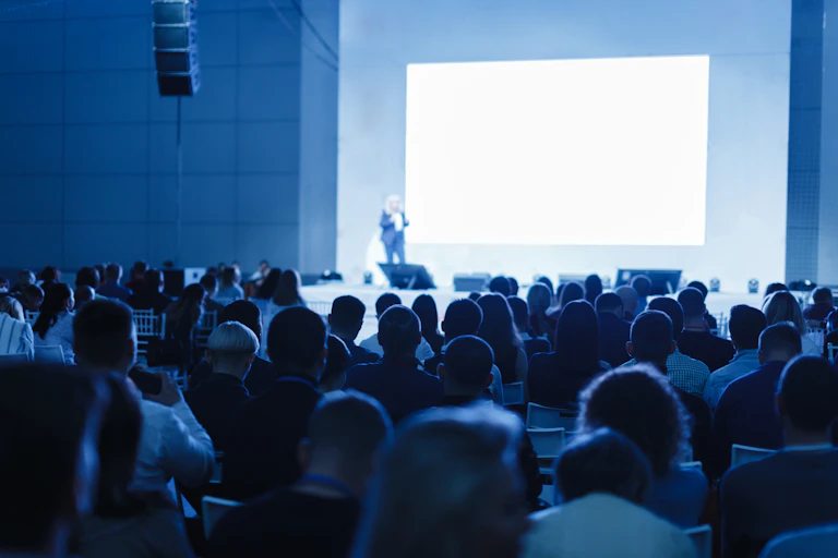 a bluish photograph of a woman presenting a to a conference to a seated audience