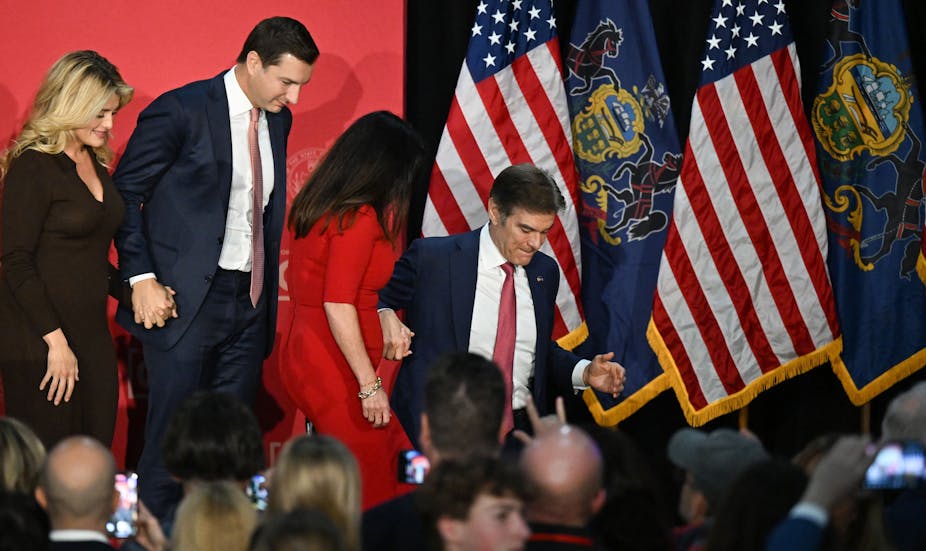 Two men in blue suits and women in formal clothing walk together as they appear to step down from a stage in front of American flags 
