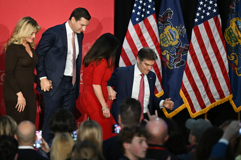 Two men in blue suits and women in formal clothing walk together as they appear to step down from a stage in front of American flags