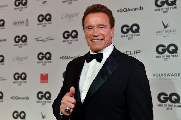 A middle aged white man with dark hair wears a tuxedo and stands in front of a white backdrop with the letters GQ on it