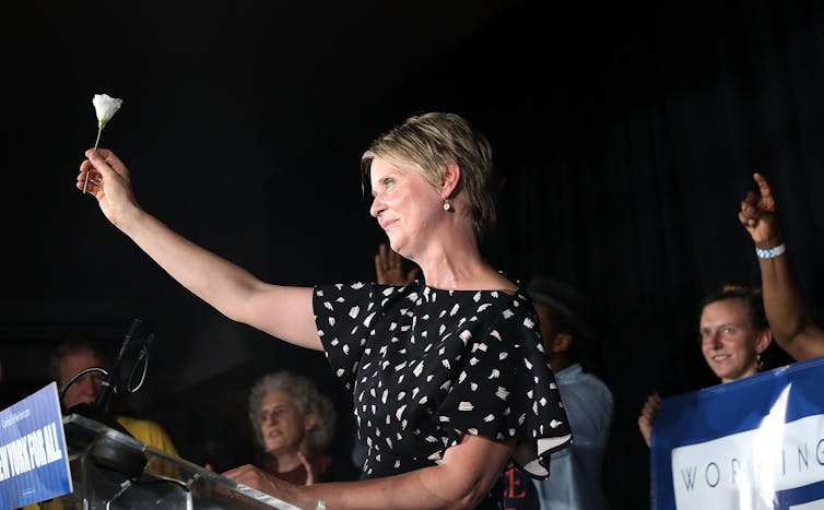 A white woman with a black shirt holds up a white rose, standing at a lectern on a dark night