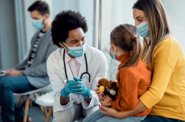 A doctor speaks to a child sitting on a woman's lap in a waiting room