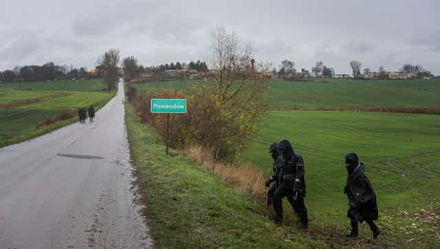 Police officers in black rain gear walk in a field and along a rural road near a road sign that reads Przewodow.