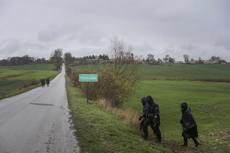 Police officers in black rain gear walk in a field and along a rural road near a road sign that reads Przewodow.