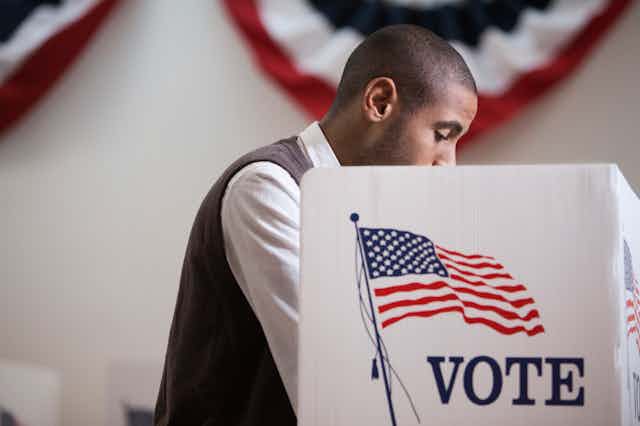 With a red, white and blue banner in the background, an Hispanic man, in a white shirt and gray vest, makes his selections at a voting booth.