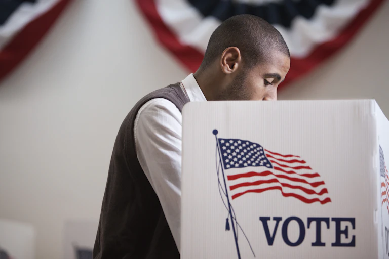 With a red, white and blue banner in the background, an Hispanic man, in a white shirt and gray vest, makes his selections at a voting booth.