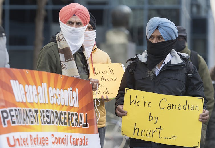 Men wearing face masks and Sikh turbans hold signs that read: we are all essential, permanent residence for all.