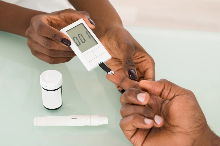 A person outside the image holds a small white biodevice in both hands so that another person - again, only their hand is pictured, can prick their index finger. A pull bottle is resting on the surface below them.