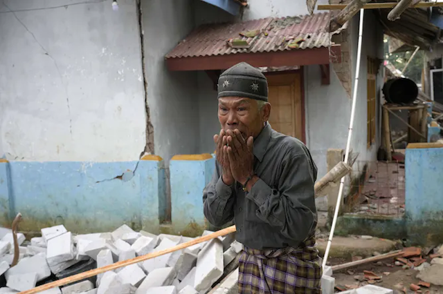Seorang laki-laki tua yang terlihat putus asa dengan reruntuhan bangunan di latar belakang