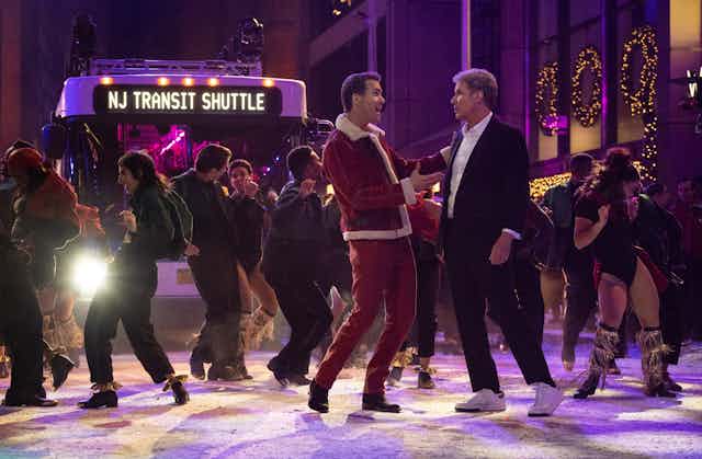 A bright bus lights dancers in the foreground, including Ryan Reynolds in a Santa outfit, and Will Ferrell in a black suit. 