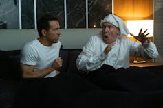 Will Ferrell and Ryan Reynolds sat in bed, Reynolds in a modern t-shirt and Ferrell in Victorian pyjamas and a Santa hat.