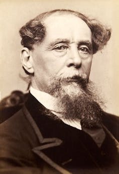A sepia portrait of a bearded Charles Dickens, posing in a black suit with velvet lapels.