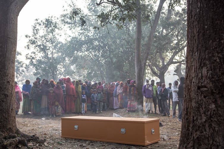 Villagers  stand around a coffin in the woods.