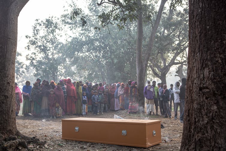 Villagers stand around a coffin in the woods.