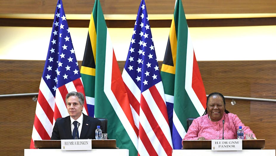 A man on a left and a woman address the media while seated. Behind them are flags of the United States and South African.