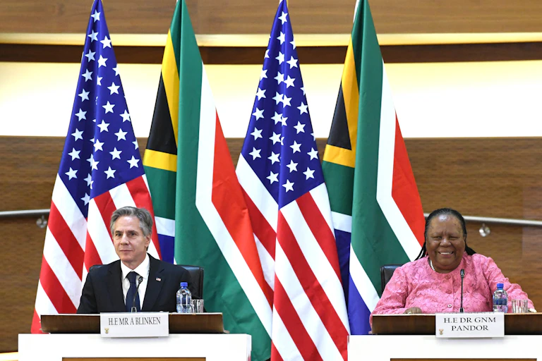 A man on a left and a woman address the media while seated. Behind them are flags of the United States and South African.
