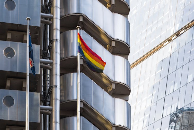 A rainbow flag on a flagpole in front of a modern silver building facade -- Lloyds building in the City of London.