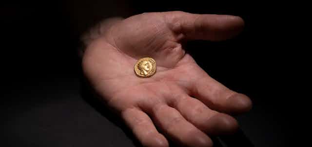 A hand holds a small gold coin in its palm featuring the head of a roman ruler. 