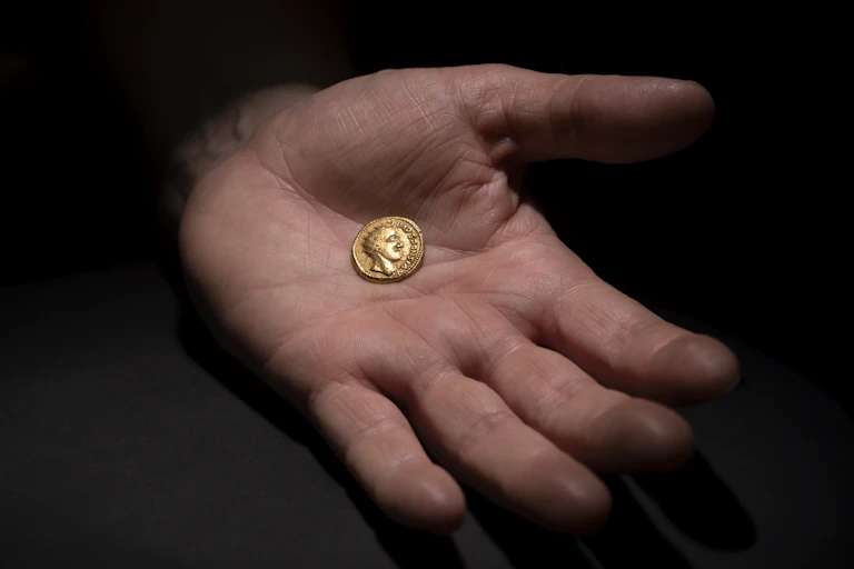 A hand holds a small gold coin in its palm featuring the head of a roman ruler.