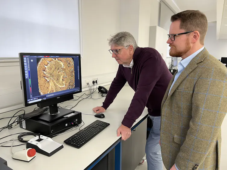Two men bend over a computer screen to look at a zoomed-in image of the coin.