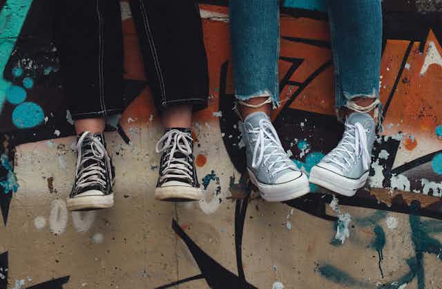 Two young people in jeans and sneakers, sitting on a wall.