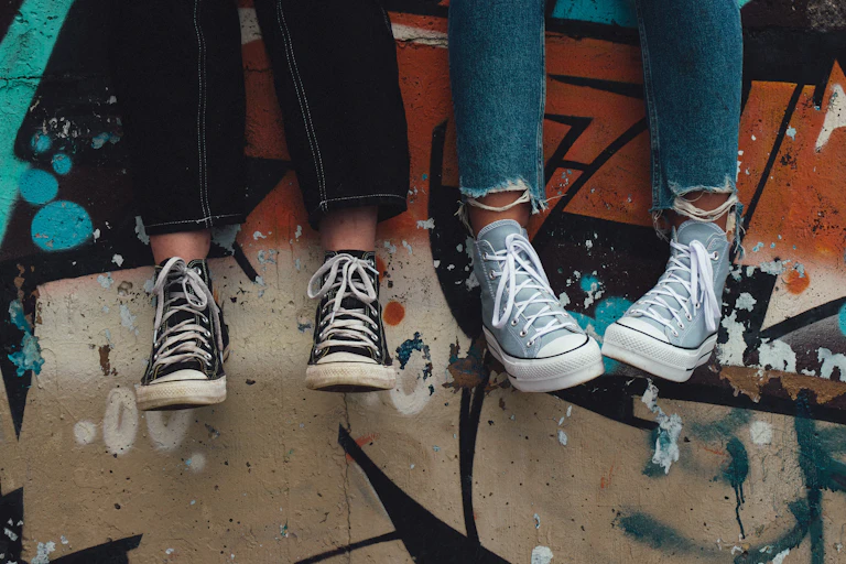 Two young people in jeans and sneakers, sitting on a wall.