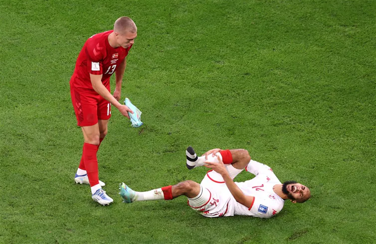 A player from the Tunisian teams lays on the pitch holding his shin in pain. A Denmark player is trying to hand him his shoe.
