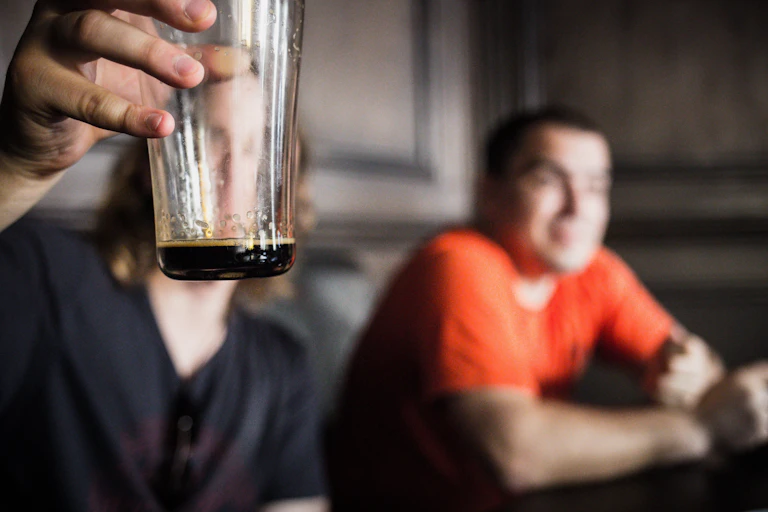 Raised, almost empty pint glass in foreground, two men in the background.
