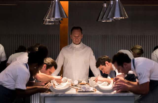 Ralph Fiennes stands at the centre of a long table of chefs, in chef whites, looking over their work. 
