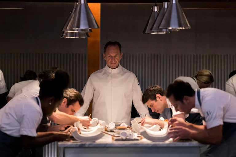 Ralph Fiennes stands at the centre of a long table of chefs, in chef whites, looking over their work.