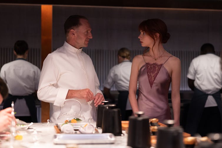Ralph Fiennes stands on the right in chef's whites, speaking to Anya Taylor-Joy, who wears a pink silk slip dress