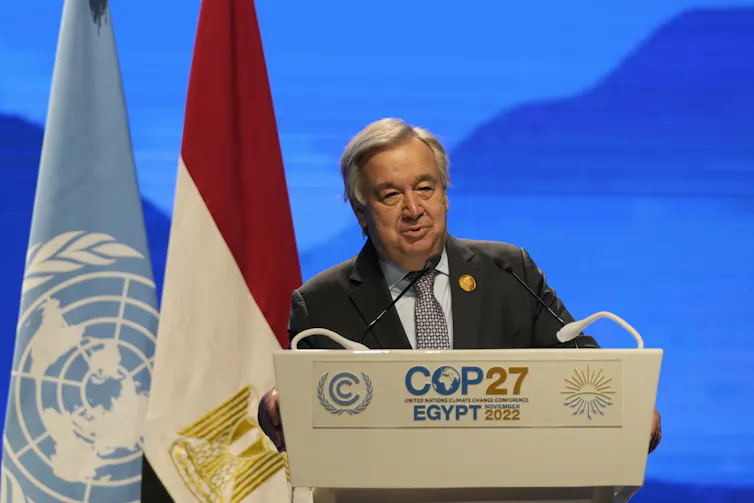 A man in a suit speaking from behind a podium that said COP27 on it. The UN flag and Egypt flag stand behind him
