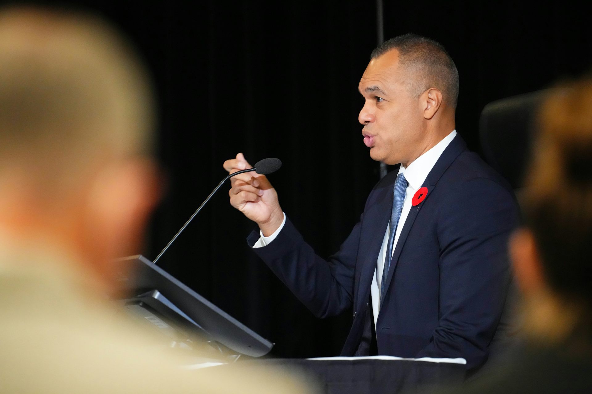 A man in a dark blue suit speaks into a microphone.