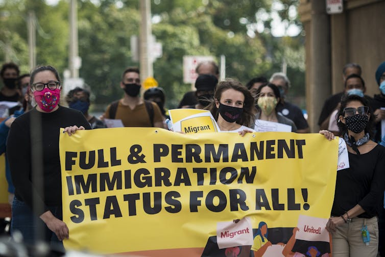 A woman wearing a face mask carries a large yellow sign with black lettering that reads: Full and permanent immigration status for all.