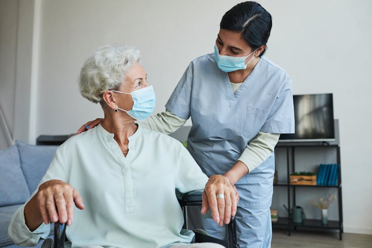 A female nurse assists a senior woman in wheelchair. Both are wearing masks