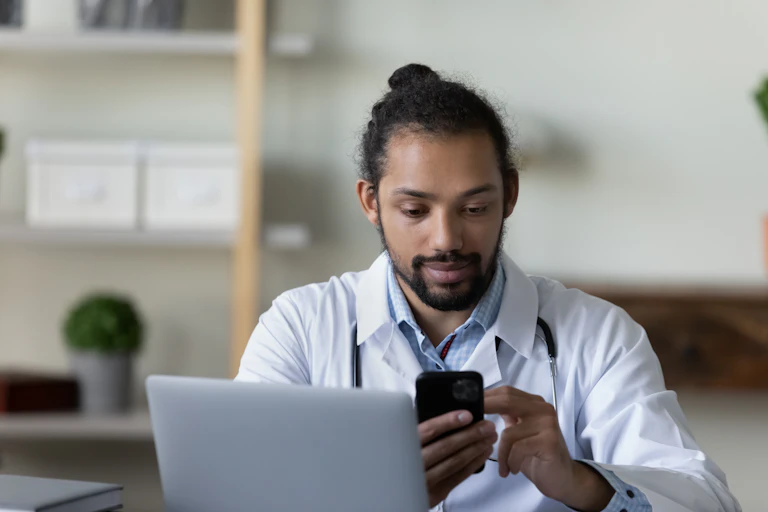 A man wearing a doctor coat sits behind a laptop, scrolling through his cellphone