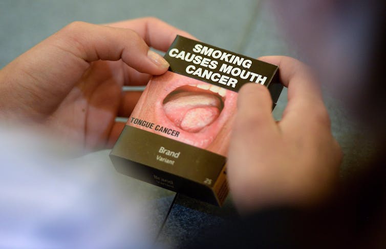 Hands holding a box of cigarettes. The box says 'Smoking Causes Mouth Cancer' with a photo of a tongue showing signs of cancer