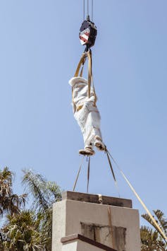 A bubble-wrapped statue is lifted into the air from its plinth by a crane.