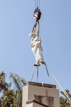 A bubble-wrapped statue is lifted into the air from its plinth by a crane.