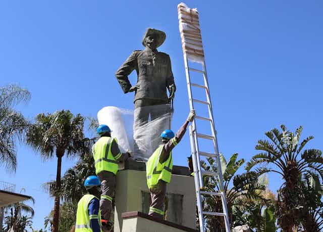 Men in yellow jackets and helmets climb the plinth of a statue of a man with a hat and an arm resting on his hip. They are wrapping the statue in bubble wrap.